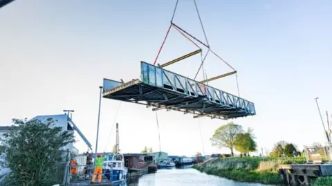 Weel Bridge being hoisted from its position on the River Hull. The bridge platform is in mid-air with a person in hi-vis clothing standing nearby. There are a number of boats moored on the waterway.