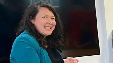 Woman with dark hair is smiling. She is wearing a black top and petrol bue jacket and is seated. 