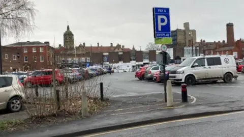 LDRS A city centre car park, filled with parked vehicles. A sign indicates a pay-and-display scheme is in operation.
