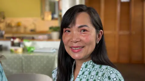 Joyce Wilmot smiles at the camera. She has long dark hair and is dressed in a smart green and white shirt. 