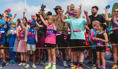 AJ Bell Young runners, and some adults, wearing running kit hold their hands aloft at the starting tape of a running event. 