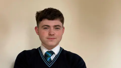 Shea. He has dark brown hair, wearing a navy school uniform.