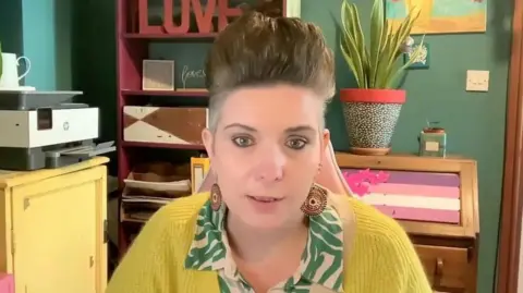 Kim Renyard A woman in a yellow jumper sat in front of a green wall.