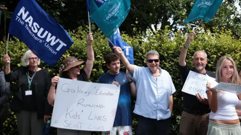 A group of six people in a row protesting. They are standing in front of a hedge and holding blue flags which either say: NASUWT or NEU. There are three men and three women. One woman in a bucket hat holds a sign saying "Galaxy Education Ruining Kid's Lives Staff lives".
