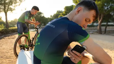 Novo Nordisk A cyclist is sitting down in the dark blue and green Team Novo Nordisk cycling gear, holding a phone to his arm, where there is a small white disc attached to the back of the upper part of his arm. A cyclist in the same outfit is leaning over his bike behind him looking at a phone. The sun is low in the sky on a sunny day and trees are around them.