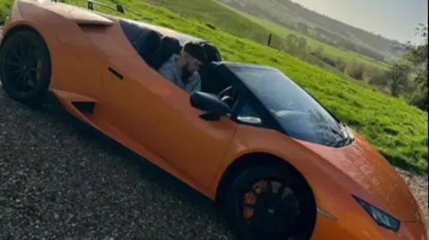 Northamptonshire Police An open-topped two seater orange sports car. Williams, with dark hair and beard, is sitting in the driver's seat. He is parked next to a field of grass. There are rolling hills in the background.