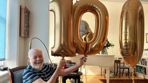 An elderly man holding gold balloons spelling out the number "100".
