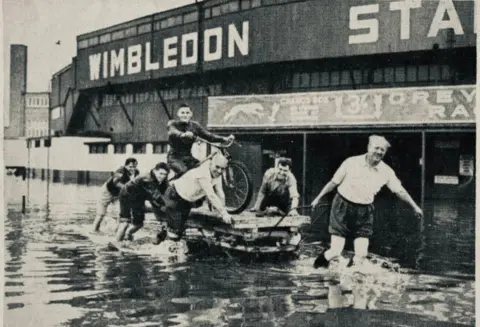 The Dons Den Ronnie Greene, the stadium promoter pulls a trolley through flood waters, Cyril Brine is mounted on the speedway bike