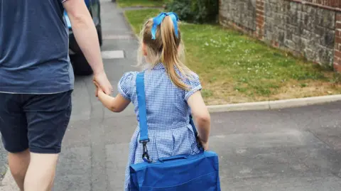 A young blonde girl in a blue gingham school dress, carrying a blue book bag with a matching ribbon in her hair. She is holding her father's hand as they walk together along the pavement beside a patch of grass and a brick wall.