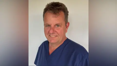 Joe Hendron A close-up of Joe in blue scrubs smiling to camera in a professional shot with a blurred white background