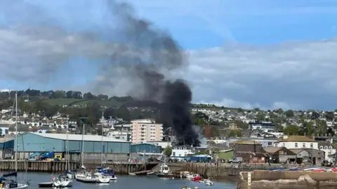 Black smoke coming from Teignmouth Library. The building is in the distance surrounded by other buildings and houses. 