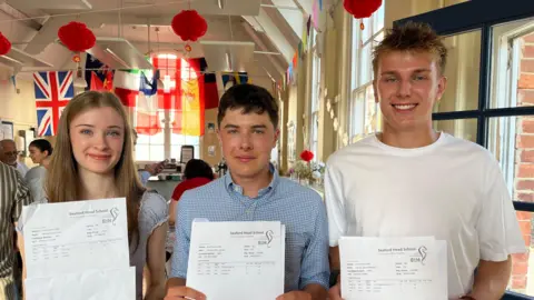 Three students stood next to each other in a row, holding pieces of paper in front of them and smiling.