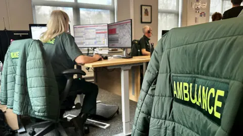 An ambulance control room with call handlers wearing headsets and a close up of a jacket over a chair with the words 'Ambulance' written on the back