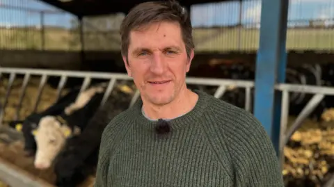 Farmer James MacCartney, a white man with brown hair, wearing a green jumper and stood in front of cows inside a farm enclosure. 