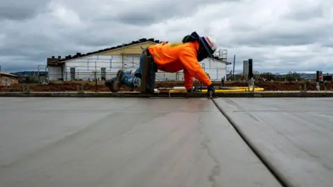 Getty Images Seorang kontraktor dengan kaus oranye terang, celana jeans dan topi konstruksi ada di tangan dan lutut yang menghaluskan lantai beton di Toll Brothers Redwood, 