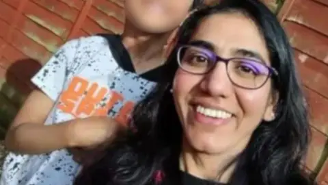 Abdhi Patel with long dark hair and glasses smiling at the camera. A child wearing a white T-shirt (whose face has been blurred) is alongside her on the left of the picture. They are both on  a red couch.