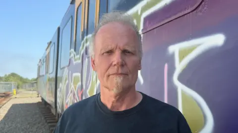 BBC/Jim Scott Martin Oliver, who is a visitor at the museum, wears a navy blue top. He is standing on the grey ballast in front of the Metro car which has been defaced with purple and green graffiti.