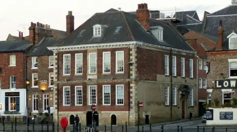 A square building with 10 white square windows on the front, next to a brickwork restaurant.