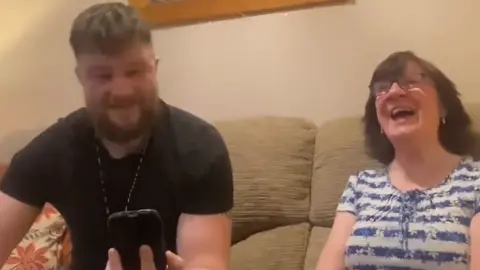 Two Connors A young man with a beard in his 20s sits with an older lady on a sofa. She is smiling and laughing and he looks proud.