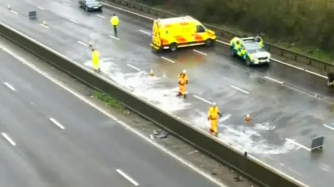 National Highways An oil spillage can be seen on a large motorway. There is a van with neon yellow and orange signage and a police car to the side. On the road are people in high vis jackets walking. 