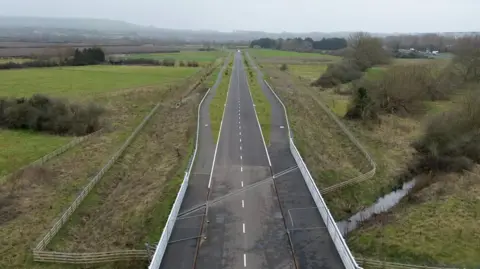 BBC An aerial shot of the unfinished and deserted road near Wanborough. There are fields either side of the Tarmac single-lane carriageway and walking paths immediately next to the road.