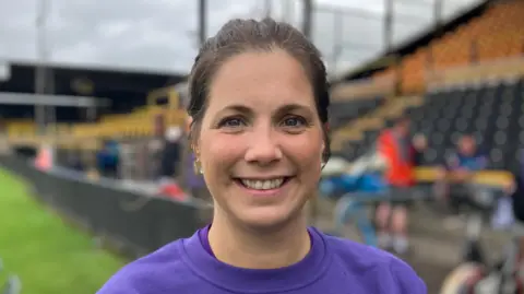 Elizabeth Baines/BBC Lindsay Burrow - a woman with tied back brown hair wearing a purple T shirt - smiles at the camera