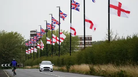 PA Media Union jack and St George's Flags hang from each of 12 lamp posts along a suburban road with a hedge along the side, with a white car and bike being ridden along the carrirageway. 