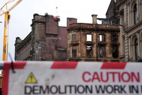 PA Media The fire-ravaged shell of a building on the corner of a street in the centre of Glasgow. There is a chimney at the top of the building, which was previously attached to the building which has now collapsed. It appears to hang in mid-air and has a red circle around it.