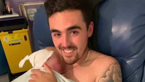 Ryan Hampson, who has black hair, is smiling and holding a newborn baby who is dressed in a white baby hat and covered in a towel.