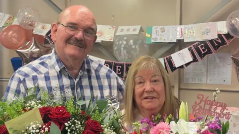Alan wearing a white and blue checkered shirt stands beside Elaine holding two bouquets of flowers. Alan is wearing glasses and has a bald head and a grey moustache. 