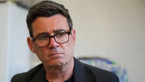 Andy Burnham, a man with dark hair and glasses. He is wearing a black suit jacket and t-shirt.