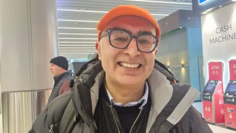 Man with orange cap and large spectacles wears a puffa jacket. Airport cash machines can be seen in the background.