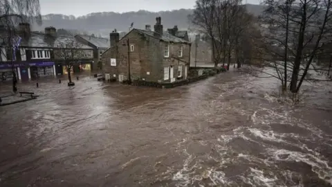 PA Media Hebden Bridge town centre under metres of flood water on Boxing Day in 2015. There are rows of stone buildings surrounded by water.