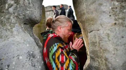 PA Media A man with long greying hair tied up in a ponytail, wearing a colourful geometric poncho. He is standing tucked between two rocks at Stonehenge with his eyes closed and his hands clasped together.