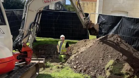 A man wearing a yellow hi-vis tabard and yellow hi-vis trousers stands in a hole which has been dug up by a digger on a patch of grass on a sunny day. The man is smiling. The digger is positioned on one side of the hole, its arm is going over the hole and its bucket is hovering above a large mound of dirt. There is construction fencing in the background and buildings behind.