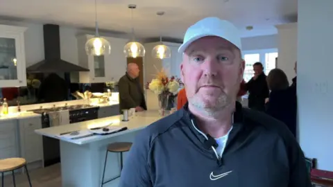 A man wearing a black quarter zip with the Nike logo on it, and a white baseball cap. He is stood in a kitchen with other people gathered in a group talking him.
