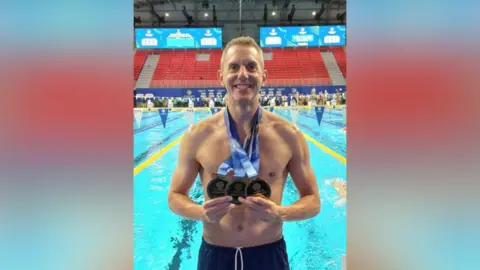 Martin Bennell A smiling man stood in front of a swimming pool holding three medals hanging around his neck