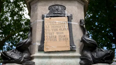 Getty Images A piece of cardboard with a message written on it reading: "This plaque is dedicated to the slaves that were taken from their homes." It was stuck on the plinth of an Edward Colston statue in Bristol after a protest in 2020.