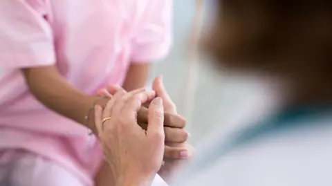 Close up of a nurse in pink scrubs holding a patient's hand.