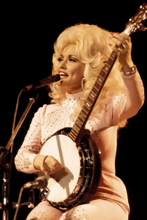 Andrew Putler/Redferns via Getty Images Dolly Parton performing live onstage at the UK Country Music Festival, playing a banjo. She wears her hair big and blond and sits before a microphone in a plae pink bedazzled shirt and pants. 