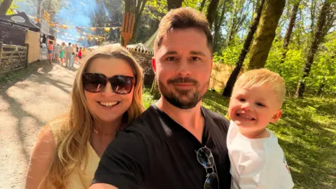 Stefan Johnson A group selfie from left to right. A woman with large sunglasses and long blonde hair is smiling next to a man with brown beard and hair weaing a black thsir with sunglasses clipped to the collar. He is holding