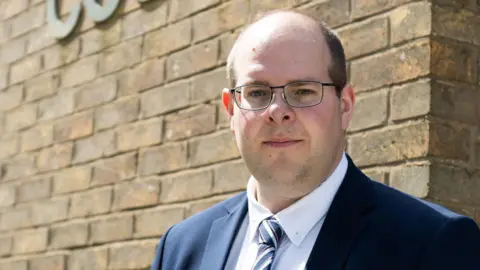 Amy Holmes/BBC Jonathan Ash-Edwards looking directly towards the camera, standing outside and in front of a brick wall. He has short hair and is bald at the front of his head. He is wearing glasses, a blue suit jacket, white shirt and striped tie. It is a bright day.