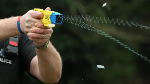 A police officer wearing black police uniform has deployed the use of a yellow taser. The taser is pointing towards the right of the picture.