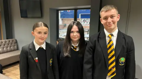 Eve, Ava and Charlie wearing their black school uniforms smiling at the camera. They are wearing black blazers, a black jumper and white shirt, and black and yellow striped ties. Eve, on the left, has brown hair. She has various badges on her blazer. Ava, in the middle, has long dark brown hair. Charlie, on the right, has shirt light brown hair.