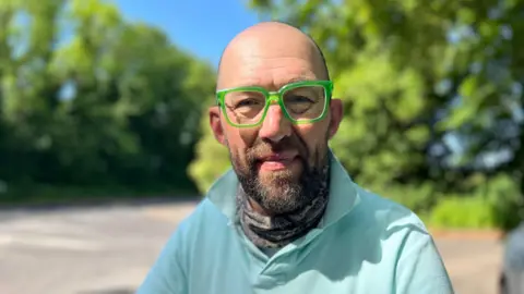George Carden/BBC Paul Kench is wearing a turquoise polo top and bright green plastic glasses. There is a road and greenery behind him. 