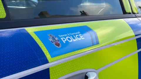 A photo of a blue sticker that reads Devon and Cornwall Police on the side of a yellow and blue police car.