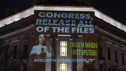 Wording is projected at nighttime onto a building that includes the phrase "Congress, release all files" in capital letters and a white font above a photo of a woman with long brown hair. The words "I was 14 when I met Epstein" are also seen in all capital letters and a green font