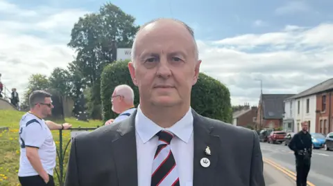 Football club chairman Darryl Cartwright with grey receding hair wearing a tie in Lostock Hall Junior FC's and suit outside the All Saints Church in Higher Walton. A couple of mourners are in the background with football tops on. It is a dry, cloudy day.
