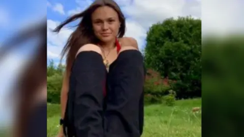PA Media Kit Birk is looking at the camera with her dark hair flowing. She is wearing black jeans and a red halterneck top.