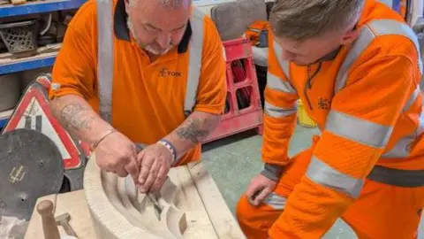 Dave Hindmarch and Josh Widdowson work on a piece of stone, wearing bright orange uniforms.